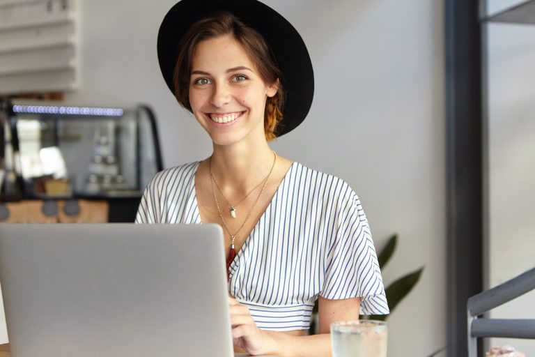 Young female freelancer in black hat and shirt sitting in office in front of opened laptop smiling rejoicing her success at work