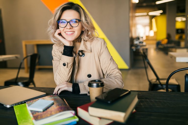 portrait of young pretty woman sitting at table in trench coat working on laptop in co-working office, wearing glasses, smiling, happy, positive, workplace