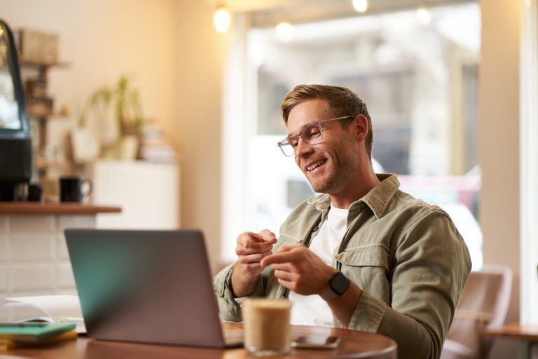 Portrait of handsome, smiling young man, online tutor, businessman working in cafe remotely, waving hand at laptop, connects to video chat or meeting