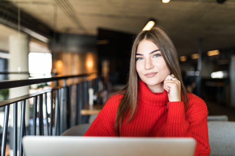 Beautiful young woman in funky hat working on laptop and smiling while sitting outdoors