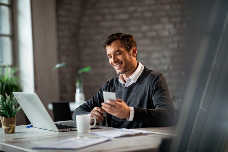 Happy businessman texting on mobile phone while working on laptop in the office