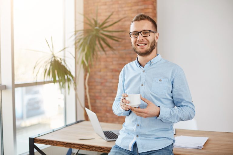 Portrait of young good-looking bearded entrepreneur in glasses and casual clothes, standing in bright coworking office, holding cup of coffee in hands, smiling in camera during interview for article in magazine