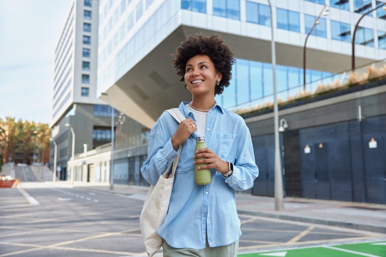 Waist up shot of happy young curly haired woman dressed in casual shirt carried fabric bag holds fresh water in bottle smiles positively strolls at city among glass skyscrapers enjoys spare time