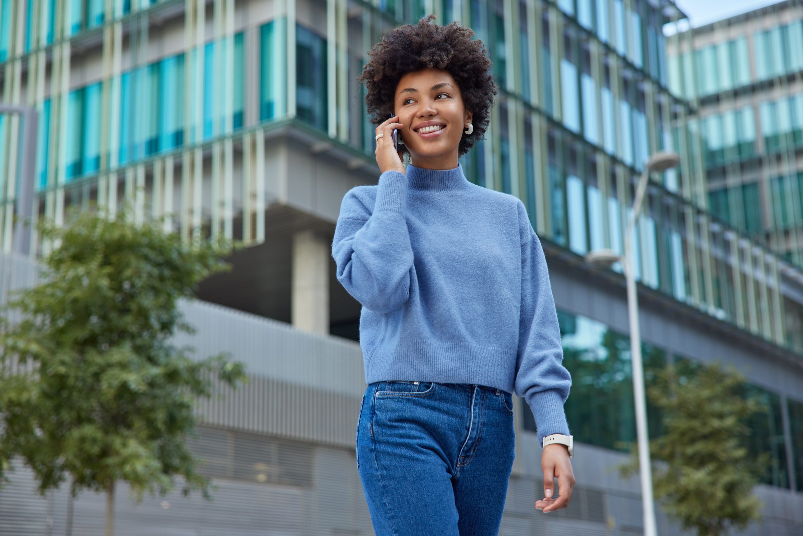 Outdoor shot of happy woman with curly hair makes call via smartphone expresses positive emotions wears casual blue jumper and jeans poses against modern glass building feels good during spare time
