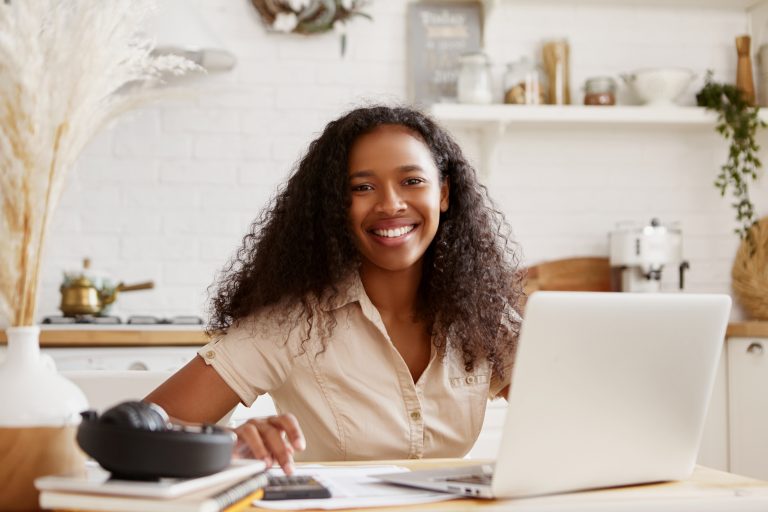 attractive-stylish-young-dark-skinned-female-beige-shirt-sitting-kitchen-table-using-laptop-calculating-budget-planning-vacations-smiling-happily-self-employed-black-woman-working-from-home