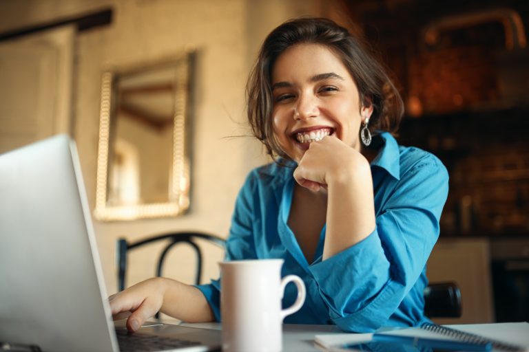 cheerful-attractive-young-woman-enjoying-distant-work-sitting-desk-using-portable-computer-drinking-coffee-pretty-female-blogger-working-from-home-uploading-video-her-channel-smiling